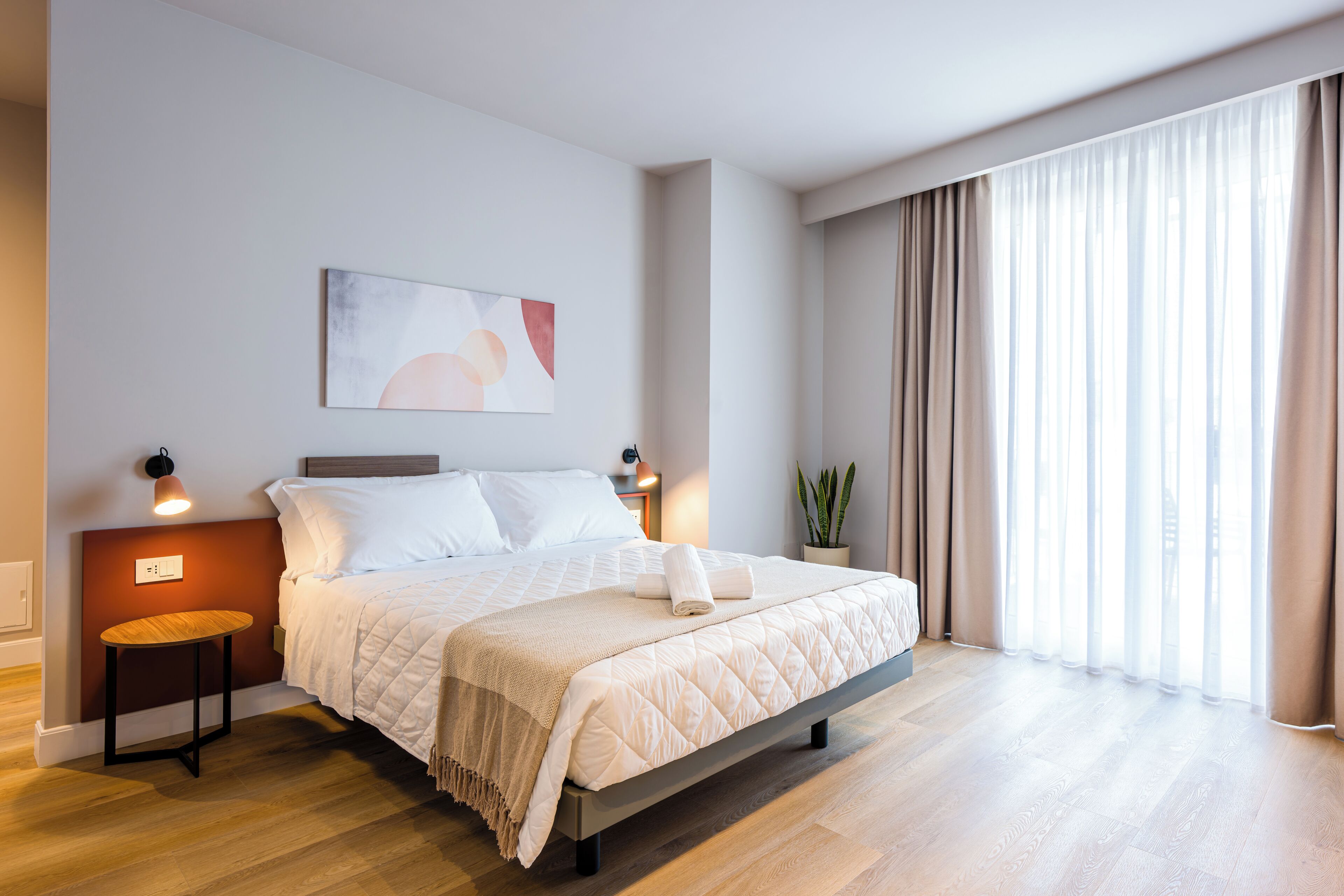 Ein modernes Hotelzimmer mit einem großen Bett, Nachttischlampen, einem Gemälde, einer Topfpflanze und Blick ins Freie.