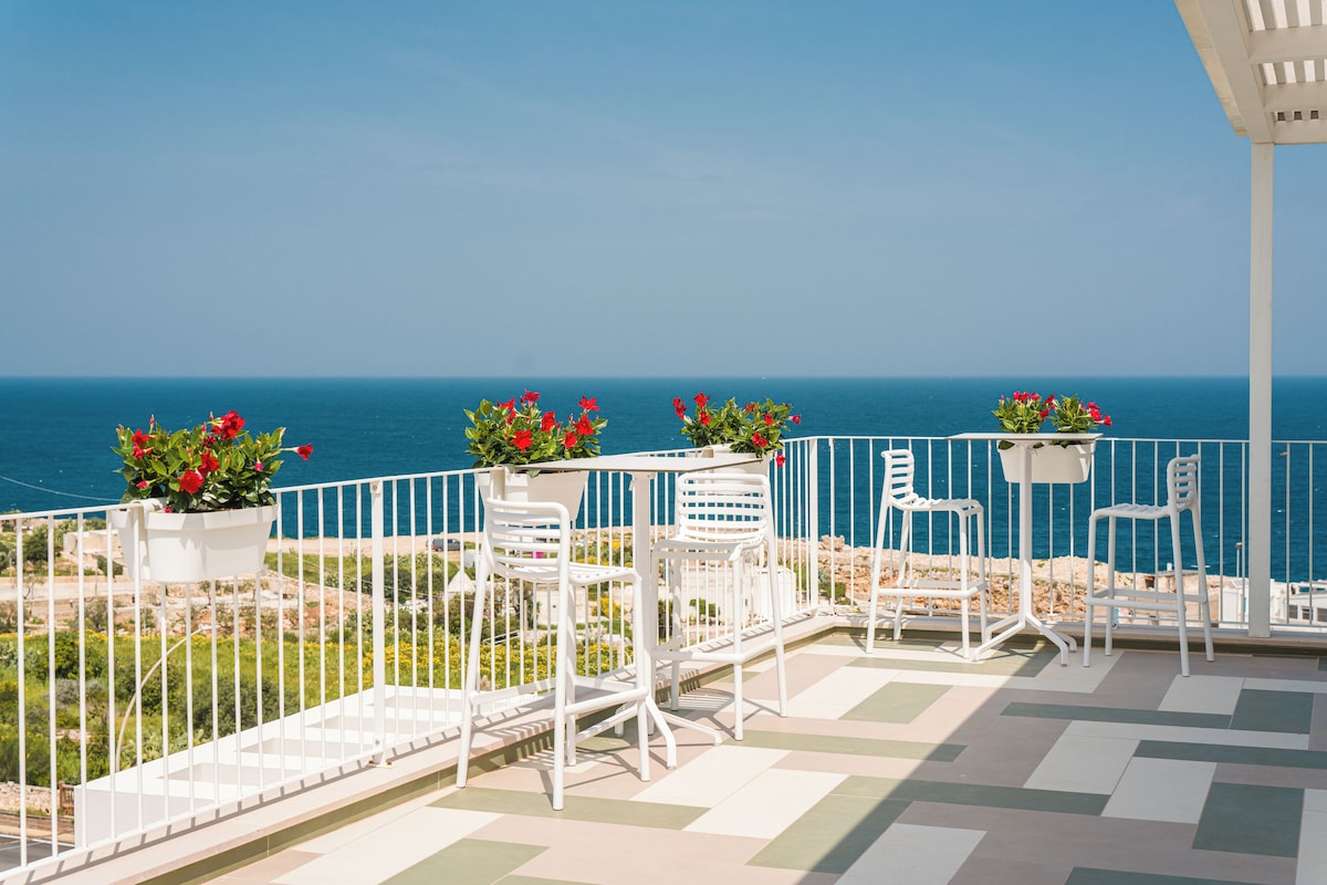 Una terrazza con sedie e tavoli bianchi, piante in vaso con fiori rossi e vista sul mare.