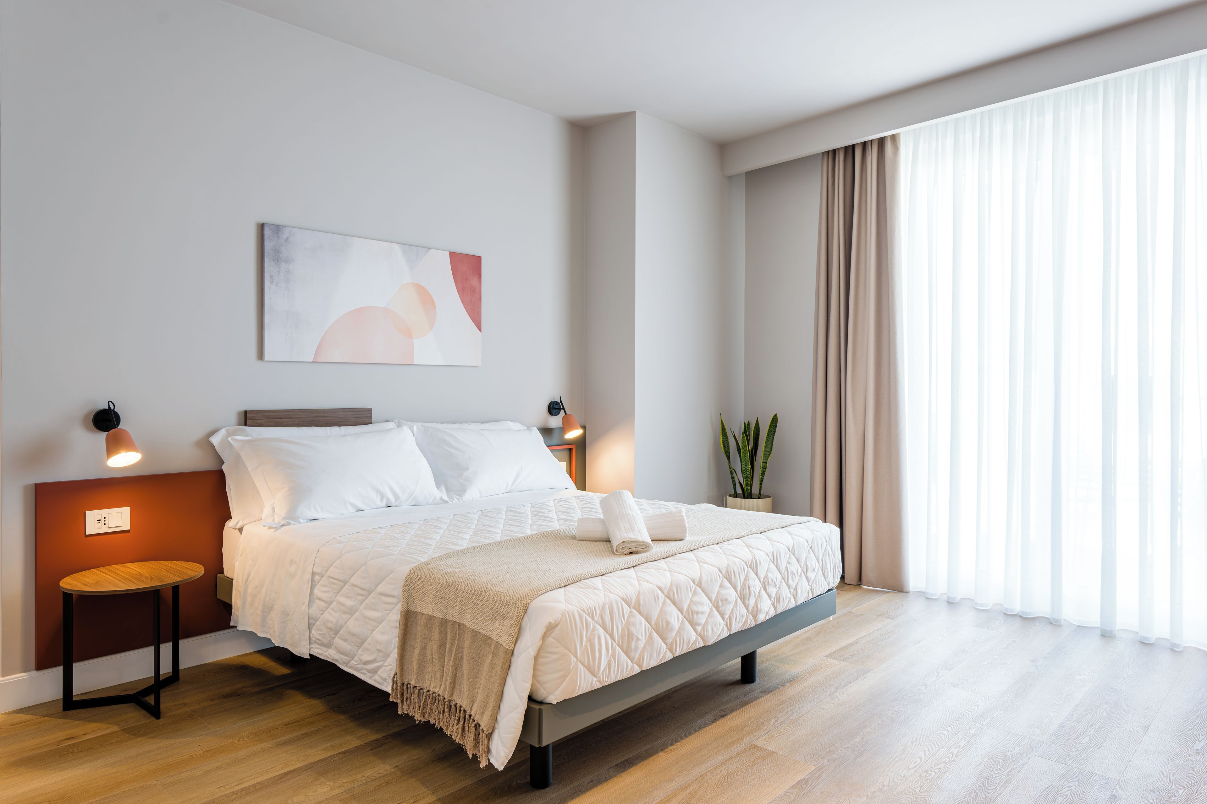 Un dormitorio moderno con una cama grande, mesitas de noche, una lámpara fijada a la pared, una planta en maceta y un ventanal con cortinas.