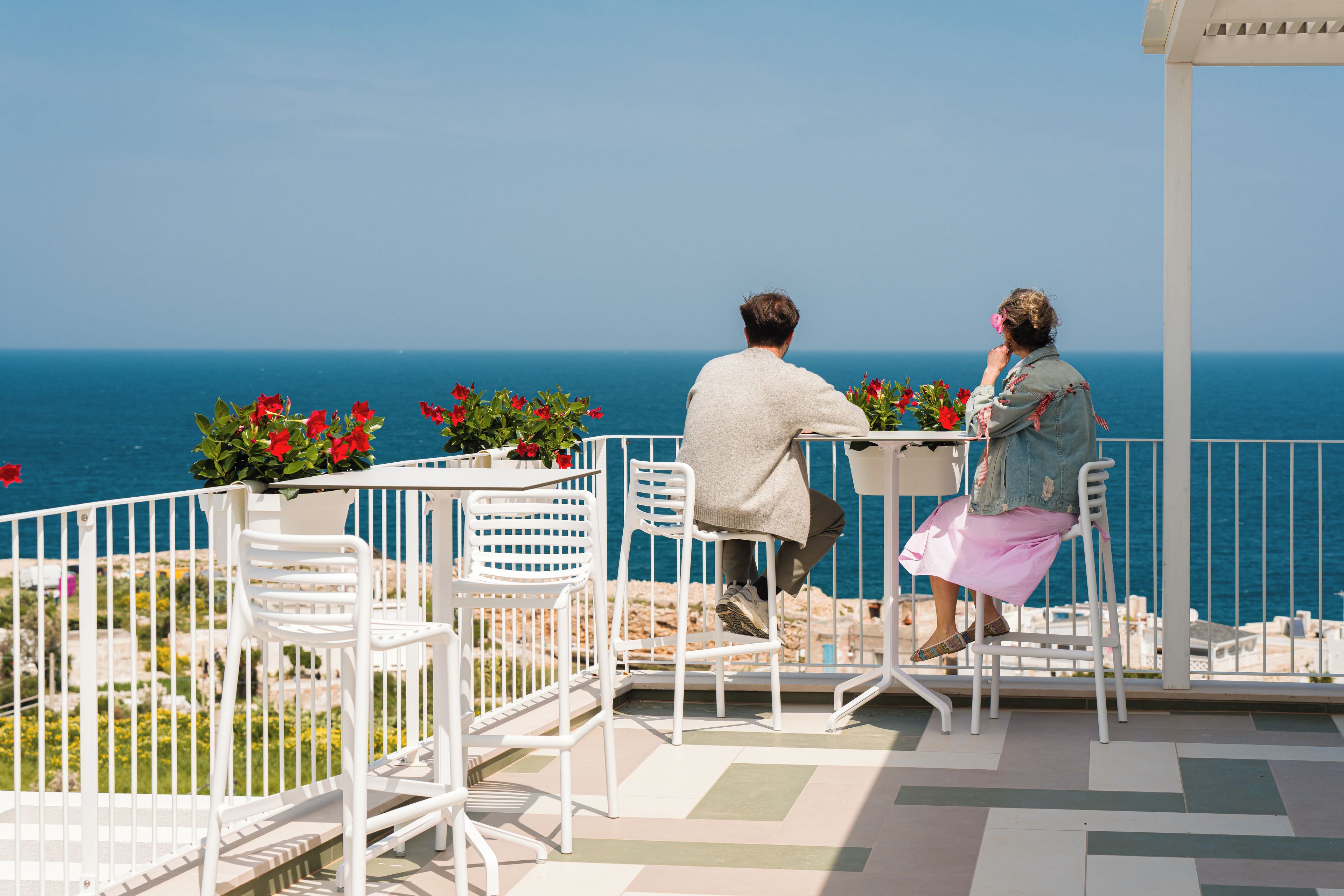 Un couple est assis à une table avec vue sur la mer, entouré de chaises blanches et de plantes en pot.