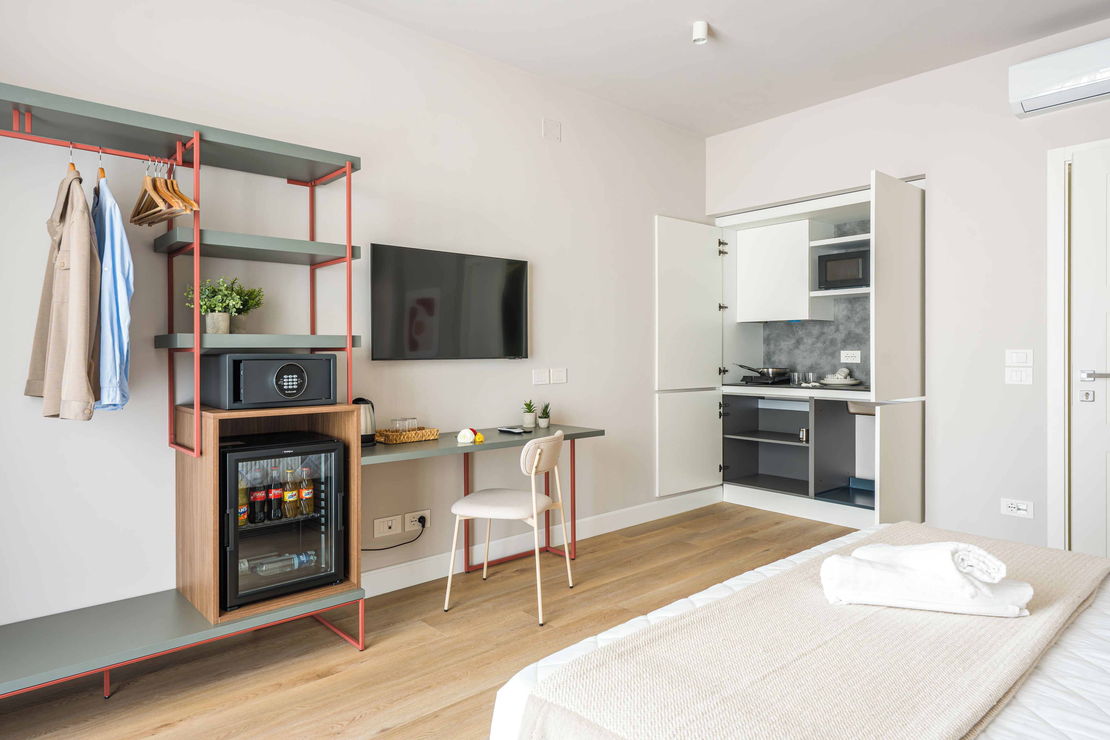 A compact hotel room with a kitchenette, a bed, a desk with a chair, and a TV mounted on the wall.
