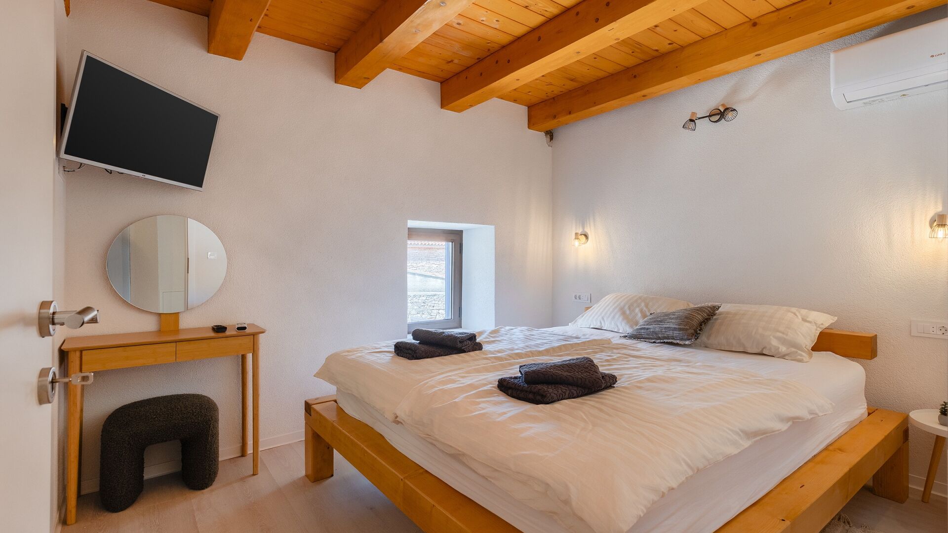 Un dormitorio con cama, mesita de noche, espejo, televisor montado en la pared y ventana.