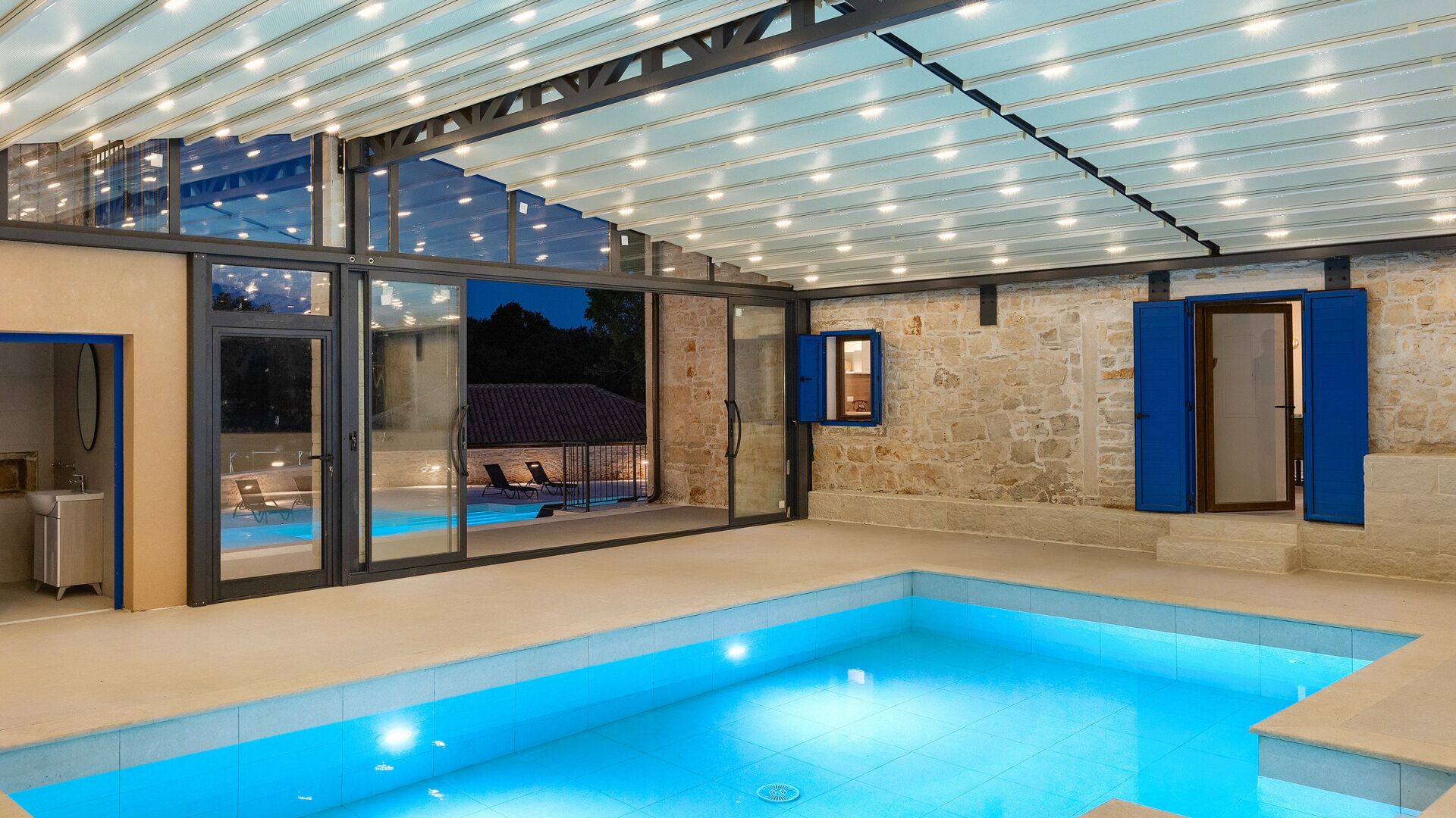 A modern indoor swimming pool with glass walls, a stone wall, and a blue door.