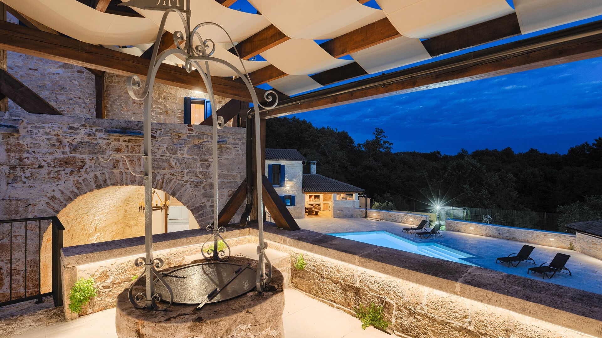 Un puits en pierre surmonté d’une structure métallique, une piscine avec des chaises longues et un bâtiment à la façade en pierre.