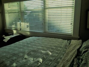 A bed with a quilt, a window with blinds, and a wall-mounted light fixture.
