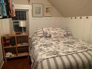 Une chambre à coucher comprenant un lit, une table de chevet, une fenêtre avec des stores et des tableaux accrochés au mur.