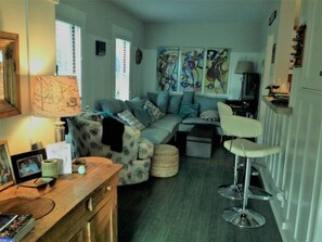 Una sala de estar acogedora con un sofá, una mesa de centro y un taburete de bar. Hay pinturas abstractas en la pared y una lámpara sobre la mesa.