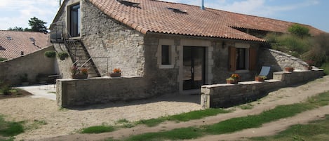A stone house with a tiled roof, a staircase leading to a balcony, and a small garden area with potted plants.