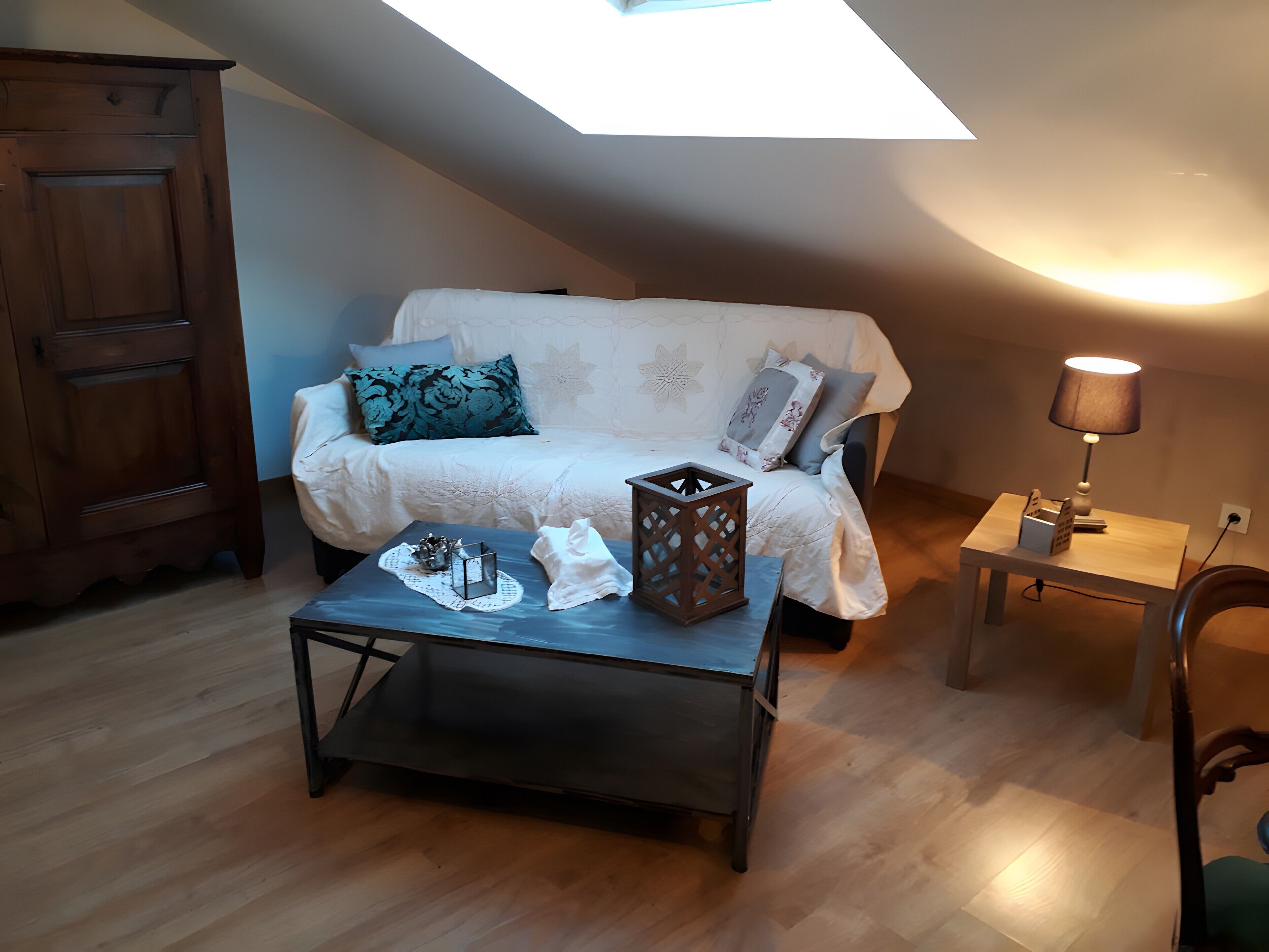 Een knus vertrek met een schuine kap, een bank met decoratieve kussens, een klein tafeltje met een lamp en een houten kast.