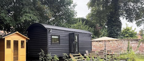 A small wooden cabin with a black exterior, a wooden shed, and a brick wall in a garden setting.