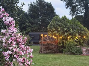 A garden with a wooden structure, possibly a gazebo, surrounded by lush greenery and flowering plants. - Cosy luxury shepherd's hut, private garden, family & pet-friendly :) (Ditchingham)