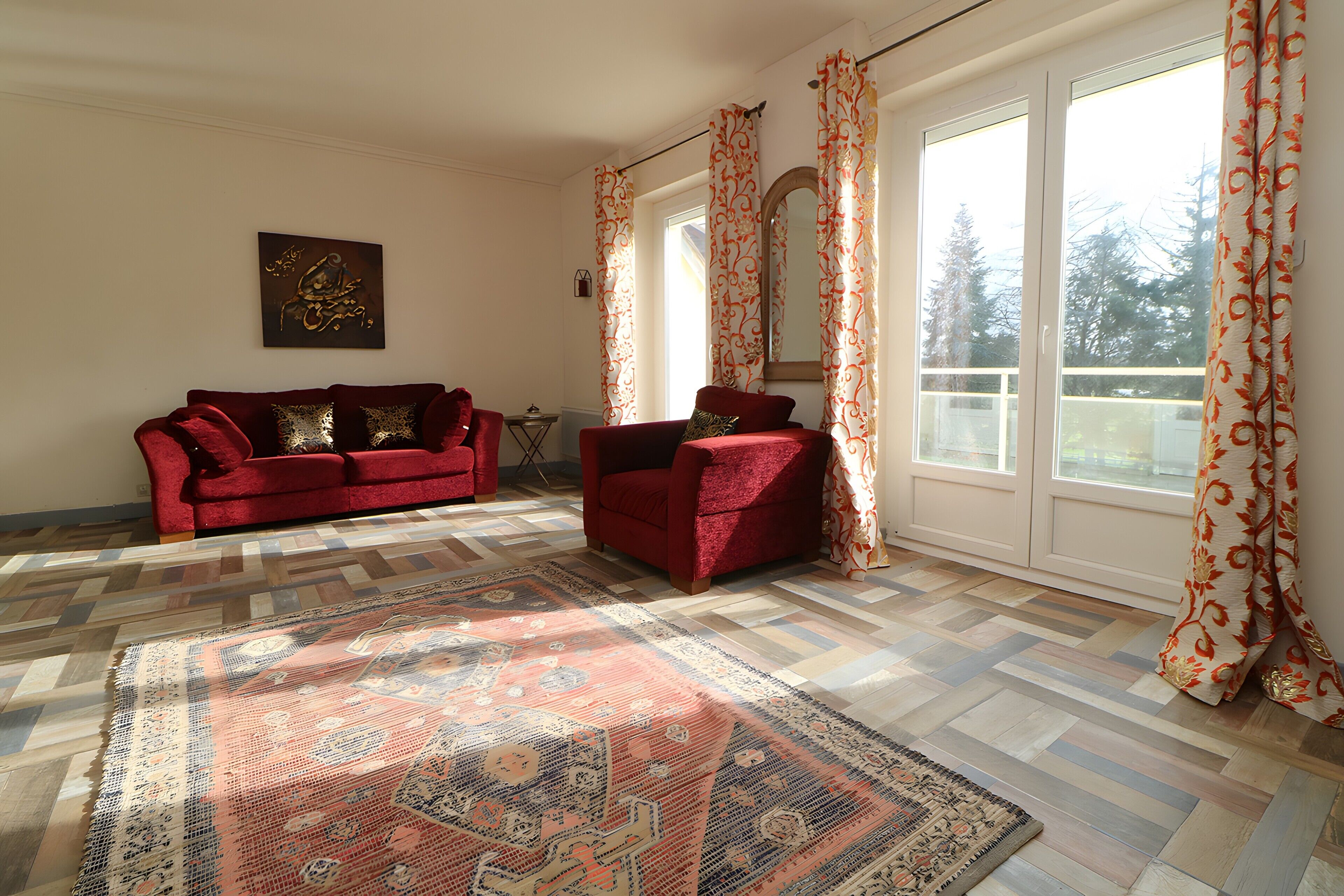 Una habitación con un sofá rojo, un tapete estampado, un espejo grande y una ventana con cortinas.