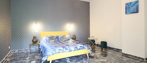 Un dormitorio con pared estampada, una cama con colcha azul y blanca, dos lámparas de noche y una lámpara moderna.
