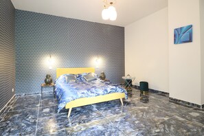 Een slaapkamer met een behang met patroon, een bed met een blauw-witte dekbedovertrek, twee nachtlampjes en een moderne lamp.