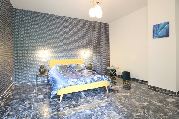 A bedroom with a patterned wall, a bed with a blue and white bedspread, two bedside lamps, and a modern light fixture.
