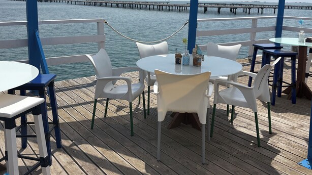 En terrasse med hvide plaststole og borde, med udsigt over vand og en anløbsbro i det fjerne.