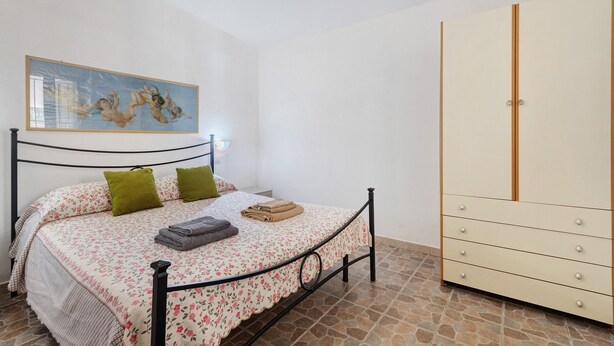 Un dormitorio con cama, almohadas, un cuadro, un armario y suelo de baldosas.