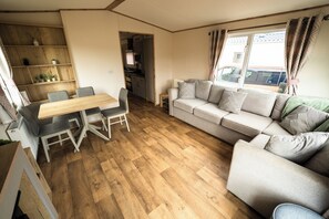 Ein Wohnzimmer mit Sofa, einem Esstisch mit Stühlen, einer Küche im Hintergrund und einem Fenster mit Vorhängen.