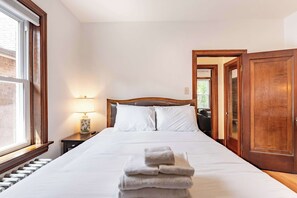 A hotel room with a bed, a nightstand with a lamp, a stack of towels, and a wooden door.