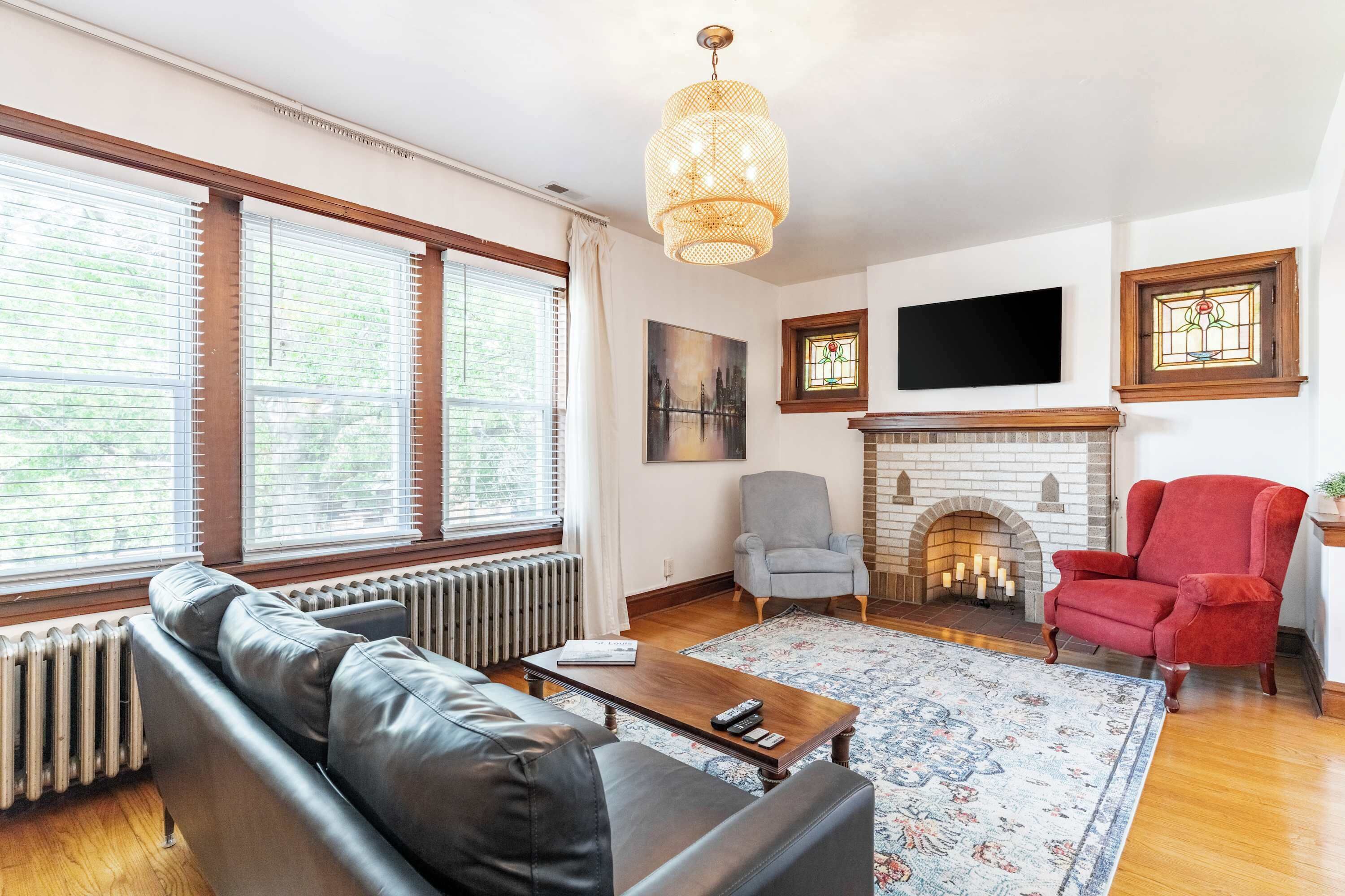 A living room with a black leather sofa, a grey armchair, a red armchair, a fireplace, and a television mounted above it.