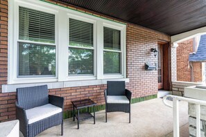 A covered patio with two wicker chairs and a bench, a brick wall, and a window with blinds.