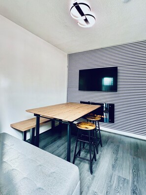 A modern dining area with a wooden table, benches, and black bar stools. A flat-screen TV is mounted on a striped wall. - Hockley 4-bed, Sleeps 12 w/ Garden & Free Parking (Birmingham)