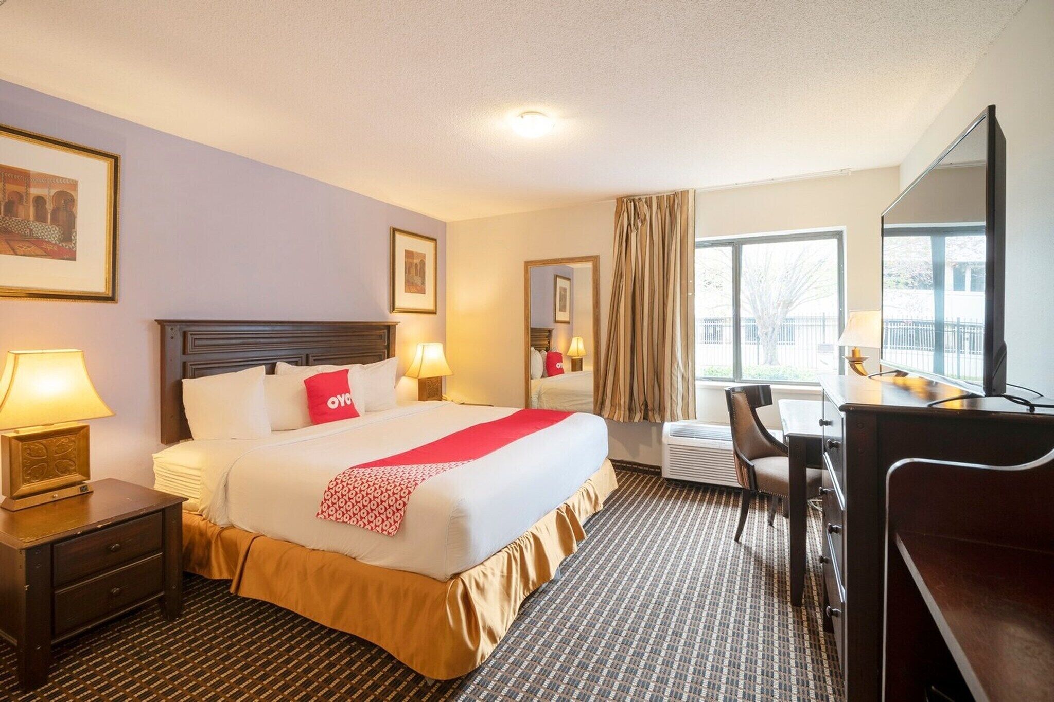 A hotel room with a bed, bedside tables, a desk, a chair, a large window, and a mirror.