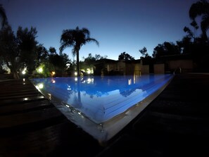 Una piscina illuminata di notte, circondata da alberi e con un pontile in legno.
