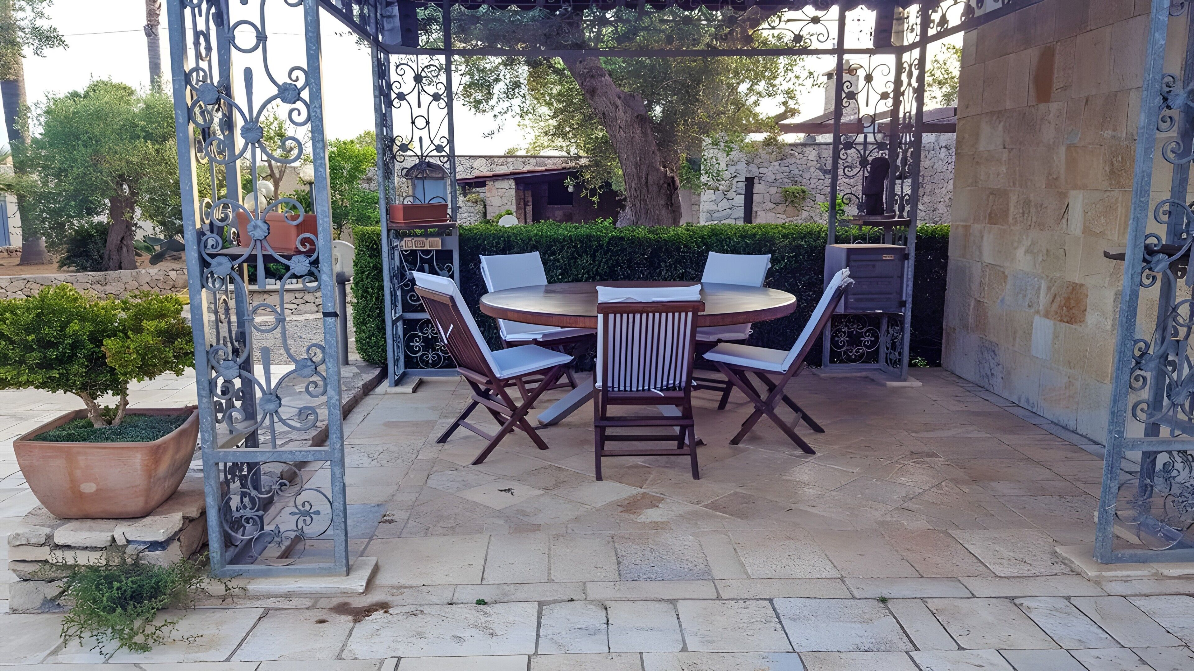 Un patio exterior con una mesa redonda y sillas, rodeado de muros de piedra y una pérgola de metal.