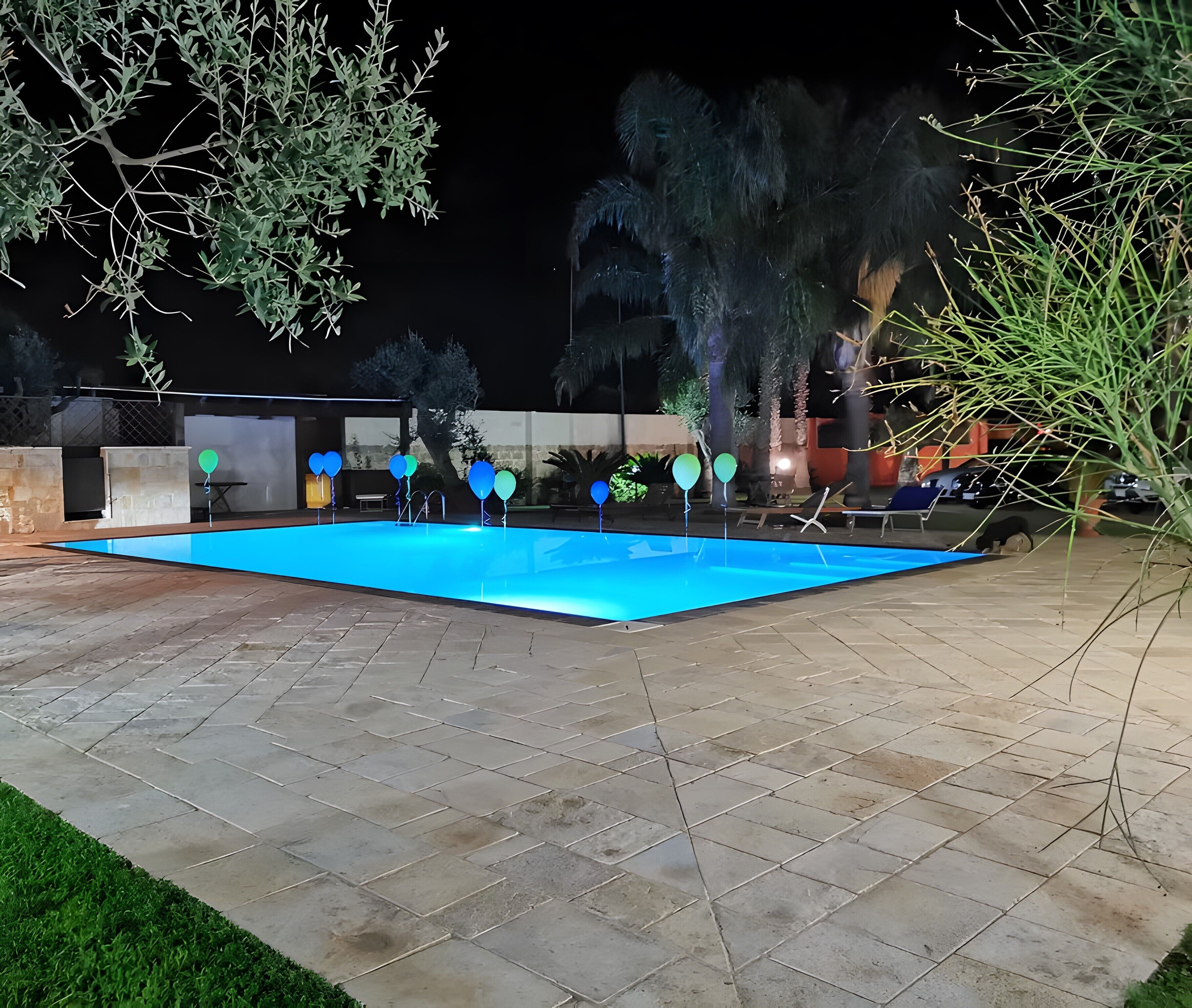 Una piscina al aire libre con solárium empedrado, rodeada de árboles y una valla.