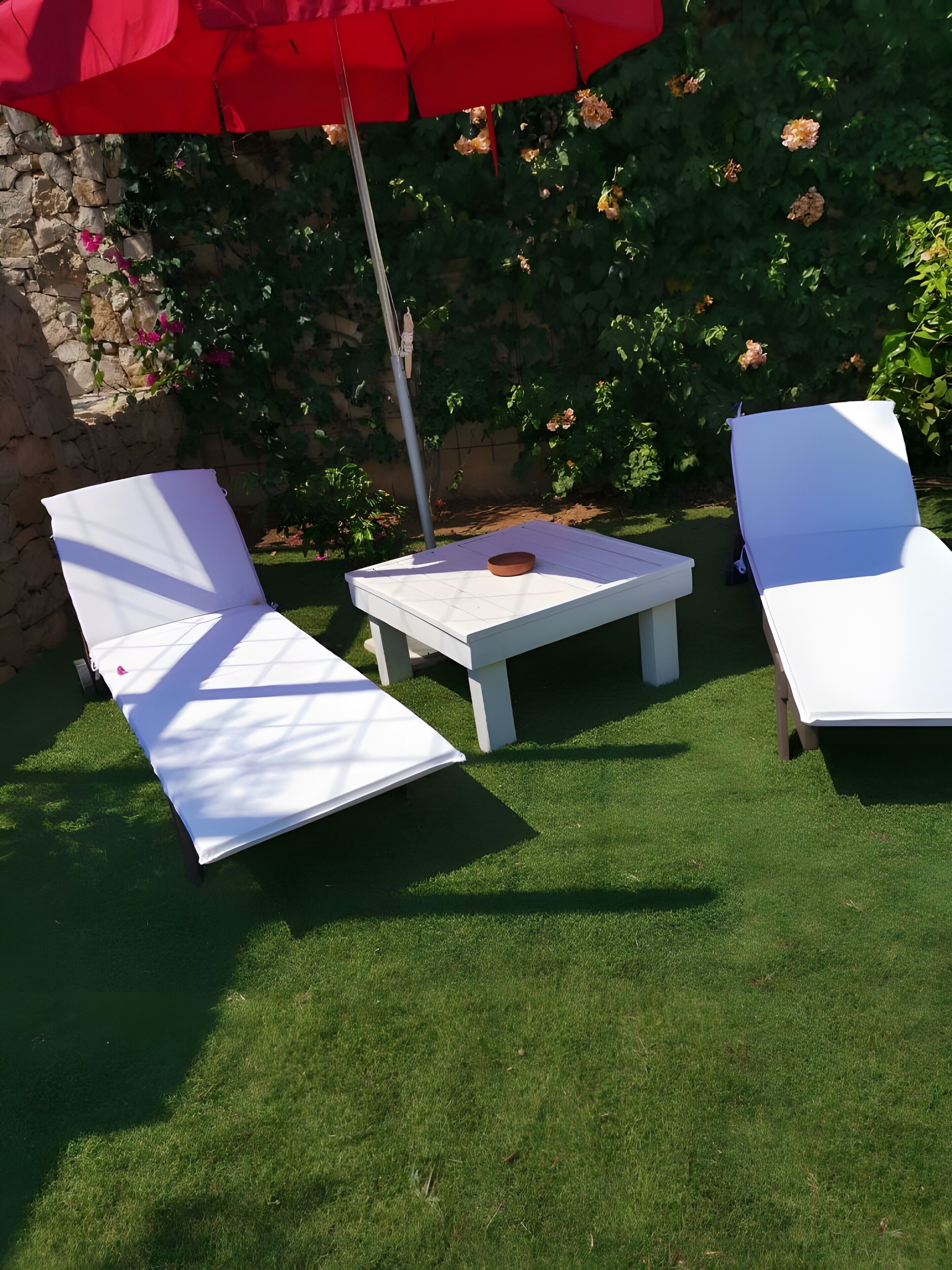 Un'area patio con due poltrone bianche e un tavolino, un ombrellone rosso e un muro di pietra con piante fiorite.