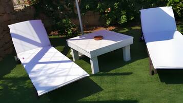 Un'area patio con due poltrone bianche e un tavolino, un ombrellone rosso e un muro di pietra con piante fiorite.