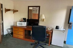 A hotel room with a wooden desk, a black office chair, a white refrigerator, a microwave, a coffee maker, a telephone, and a wall-mounted lamp.