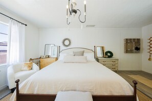 Ein ordentlich eingerichtetes Schlafzimmer mit einem großen Bett, einem Nachttisch, einem Sessel, einer Kommode und einer Kandelaber.