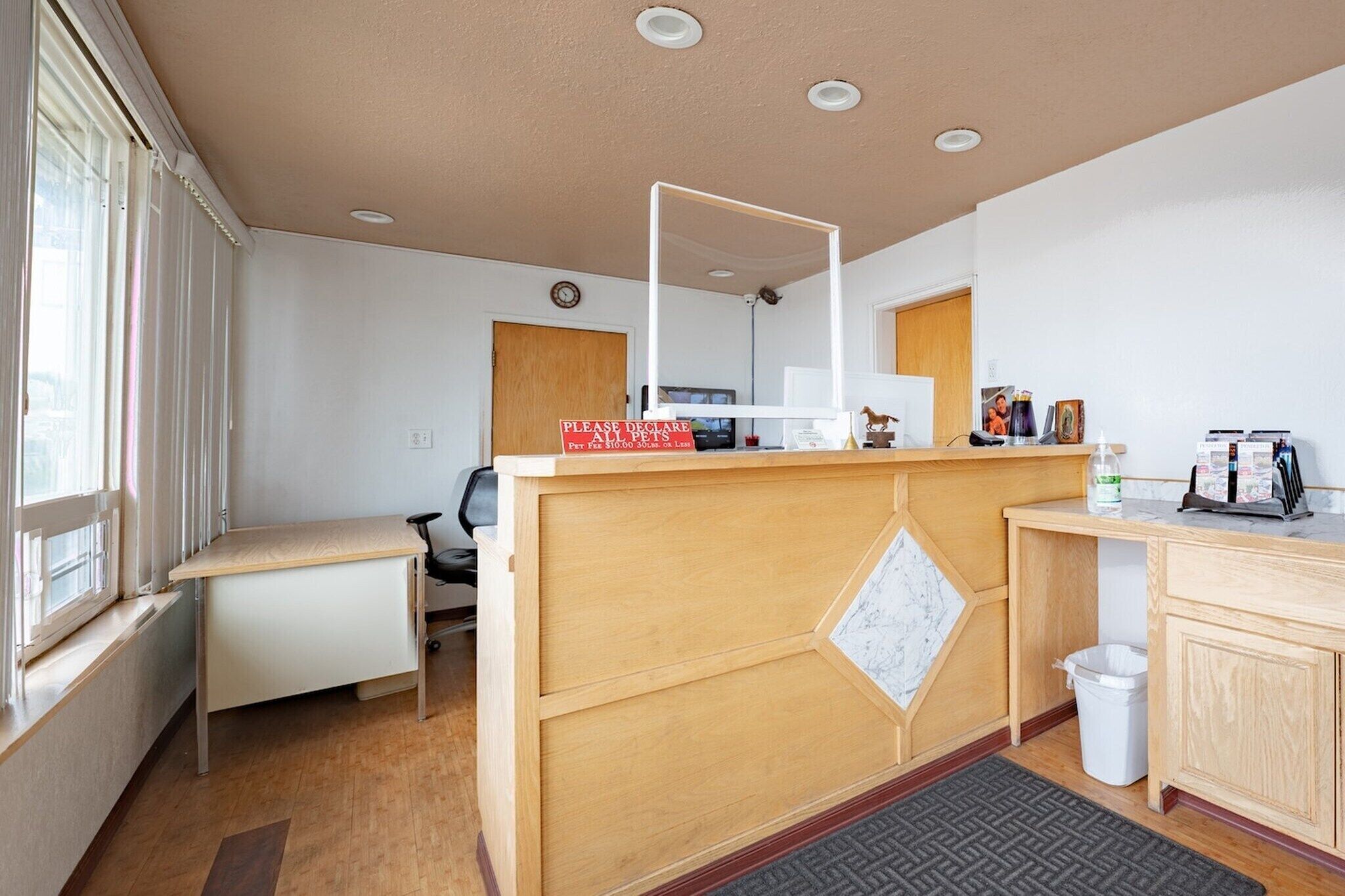 A hotel room with a wooden desk, a small chair, a mirror, a radiator, a trash bin, and a window with curtains.