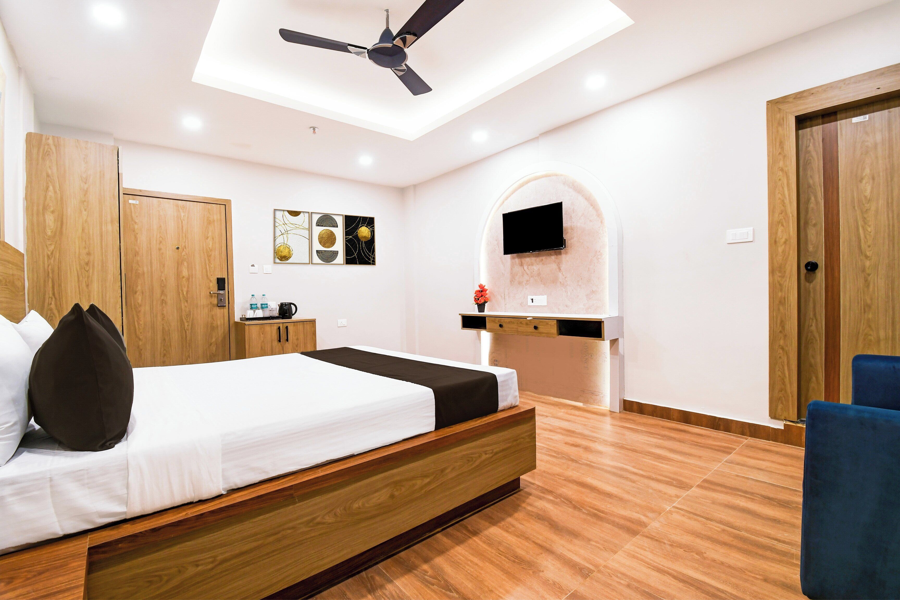 Una habitación de hotel moderna con una cama de madera, ventilador de techo, televisor montado y una kitchenette pequeña.