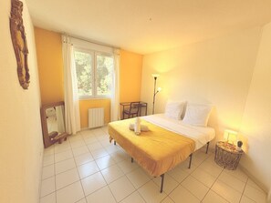 Quarto de hotel com cama, poltrona, mesa, luminária e janela com cortinas.