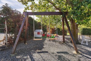 A playground with a swing set, a small playhouse, and a gravel surface. - Apartment 'Agriturismo Case Nuove - Sole' with Shared Pool, Wi-Fi and Air Conditioning (Castellina Marittima)