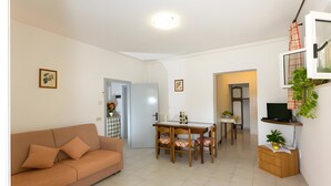Ein Wohnzimmer mit einer Couch, einem Esstisch mit Stühlen, einem Fernseher auf einem Schrank und einer Küchenzeile im Hintergrund.