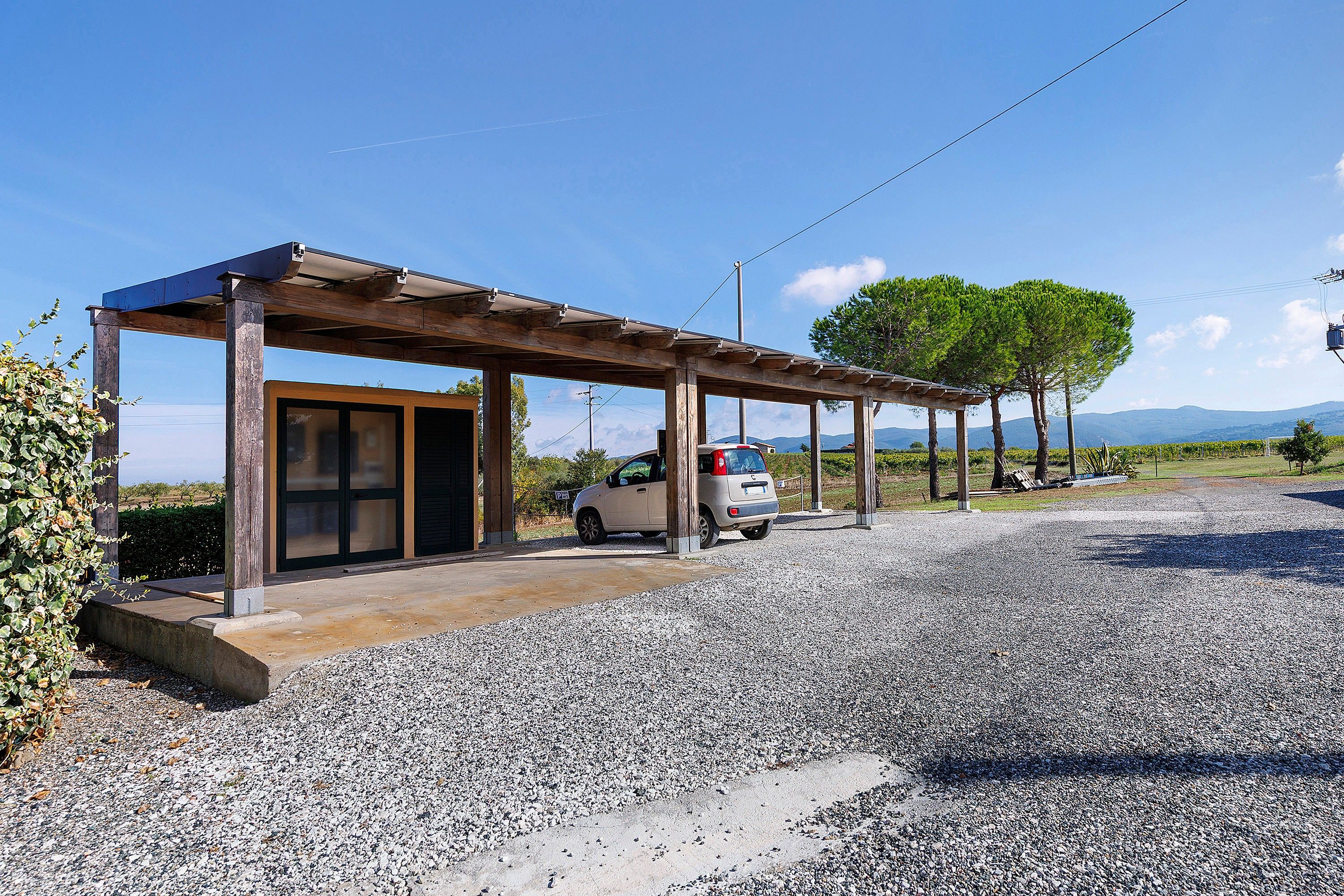 A covered outdoor area with a car parked under a pergola, gravel ground, and a clear sky.