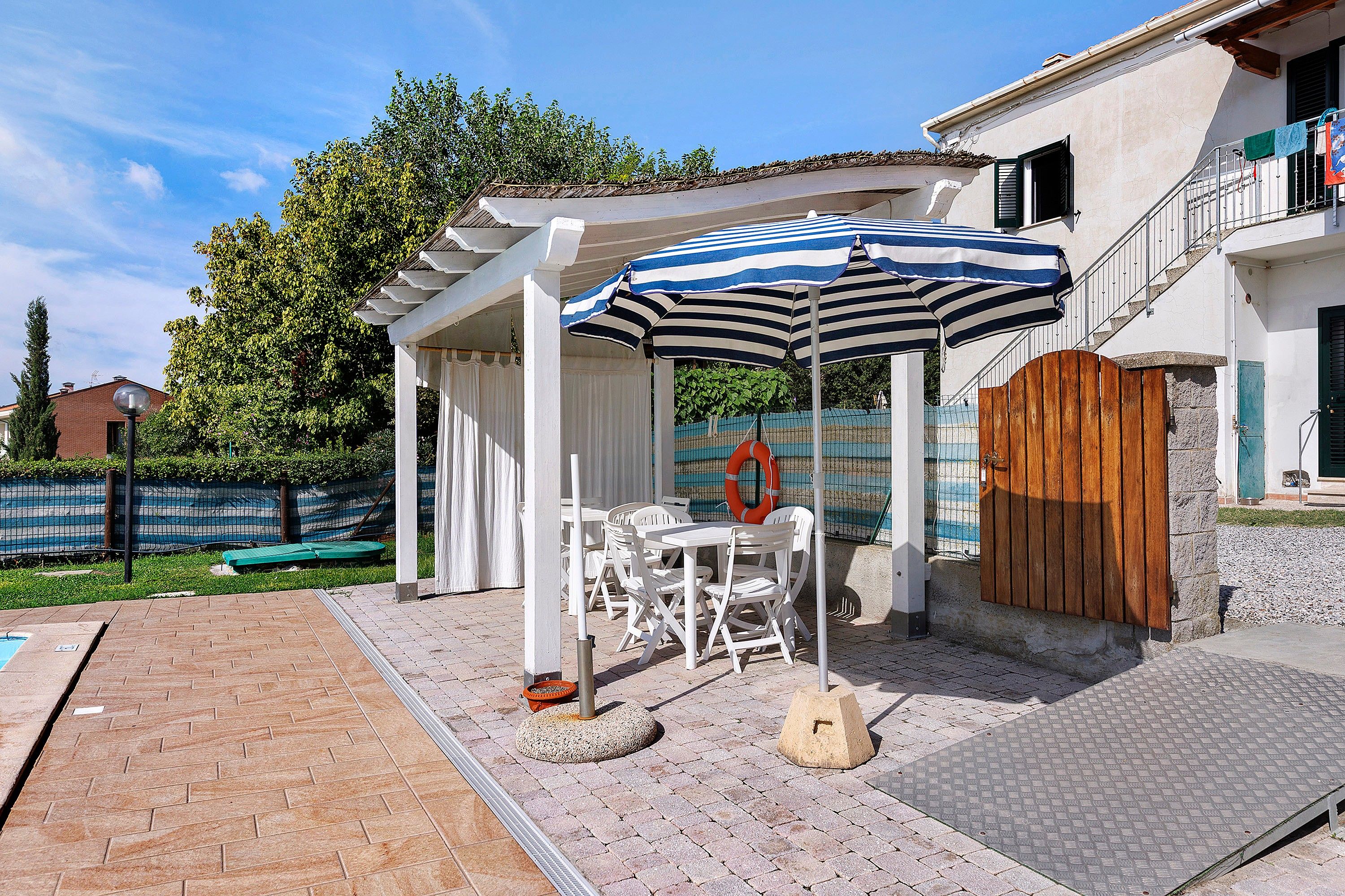 A covered outdoor seating area with a table and chairs, a striped umbrella, and a wooden gate.
