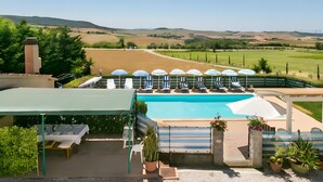 A pool area with lounge chairs and umbrellas, surrounded by a fence, with a view of the countryside. - Apartment 'Agriturismo Case Nuove - Girasole' with Shared Pool, Wi-Fi and Air Conditioning (Castellina Marittima)