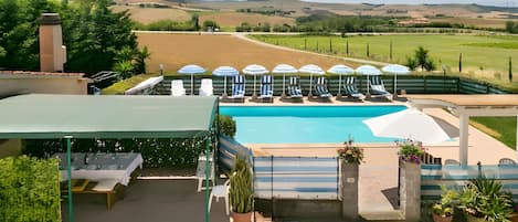 Zona de piscina con sillas de descanso y sombrillas, rodeada por una valla y con vistas al campo.