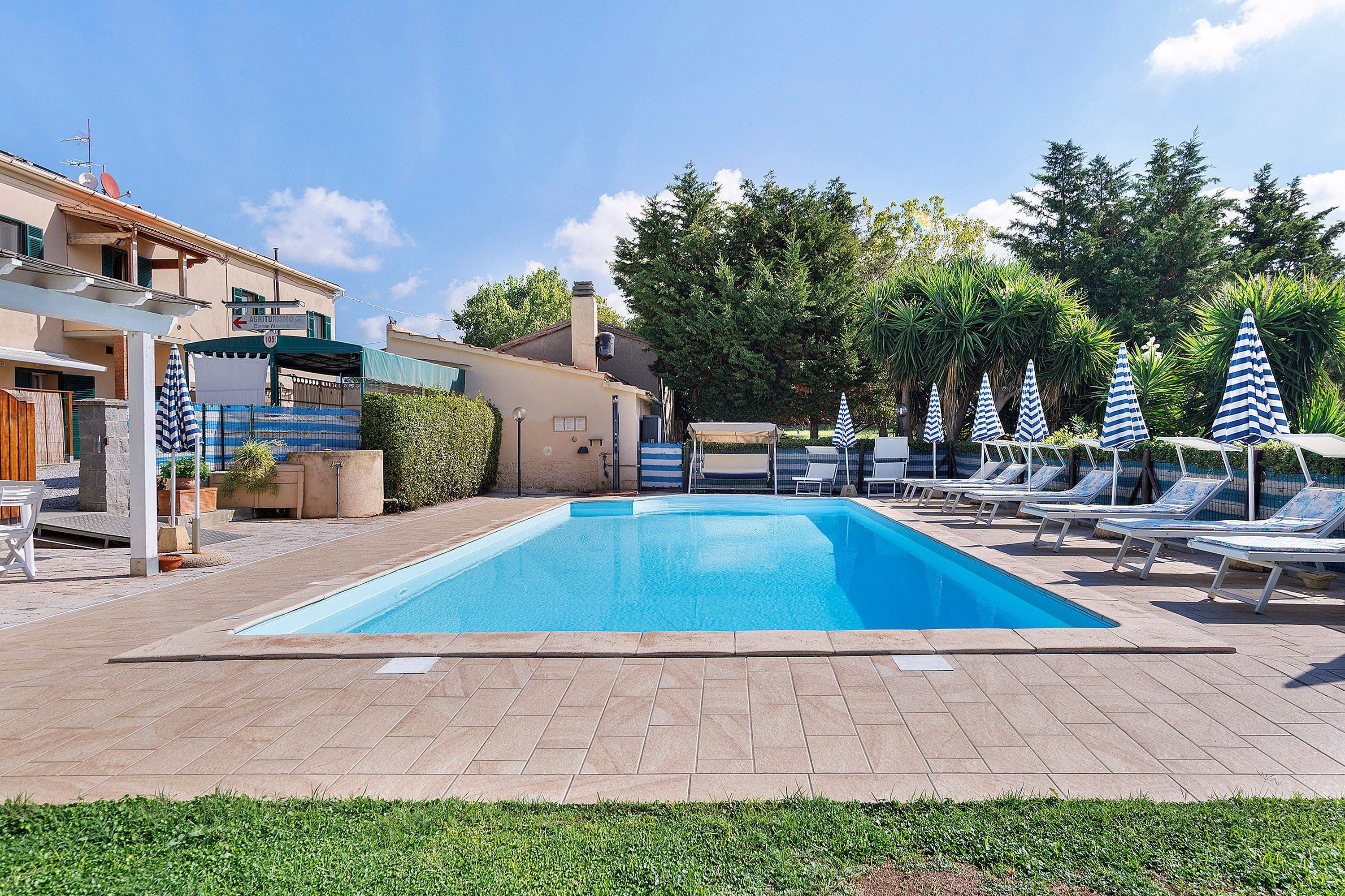 A swimming pool with lounge chairs and umbrellas, surrounded by buildings and greenery.