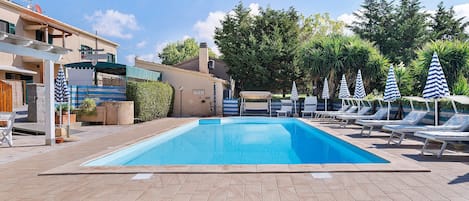 A swimming pool with lounge chairs and umbrellas, surrounded by buildings and greenery.