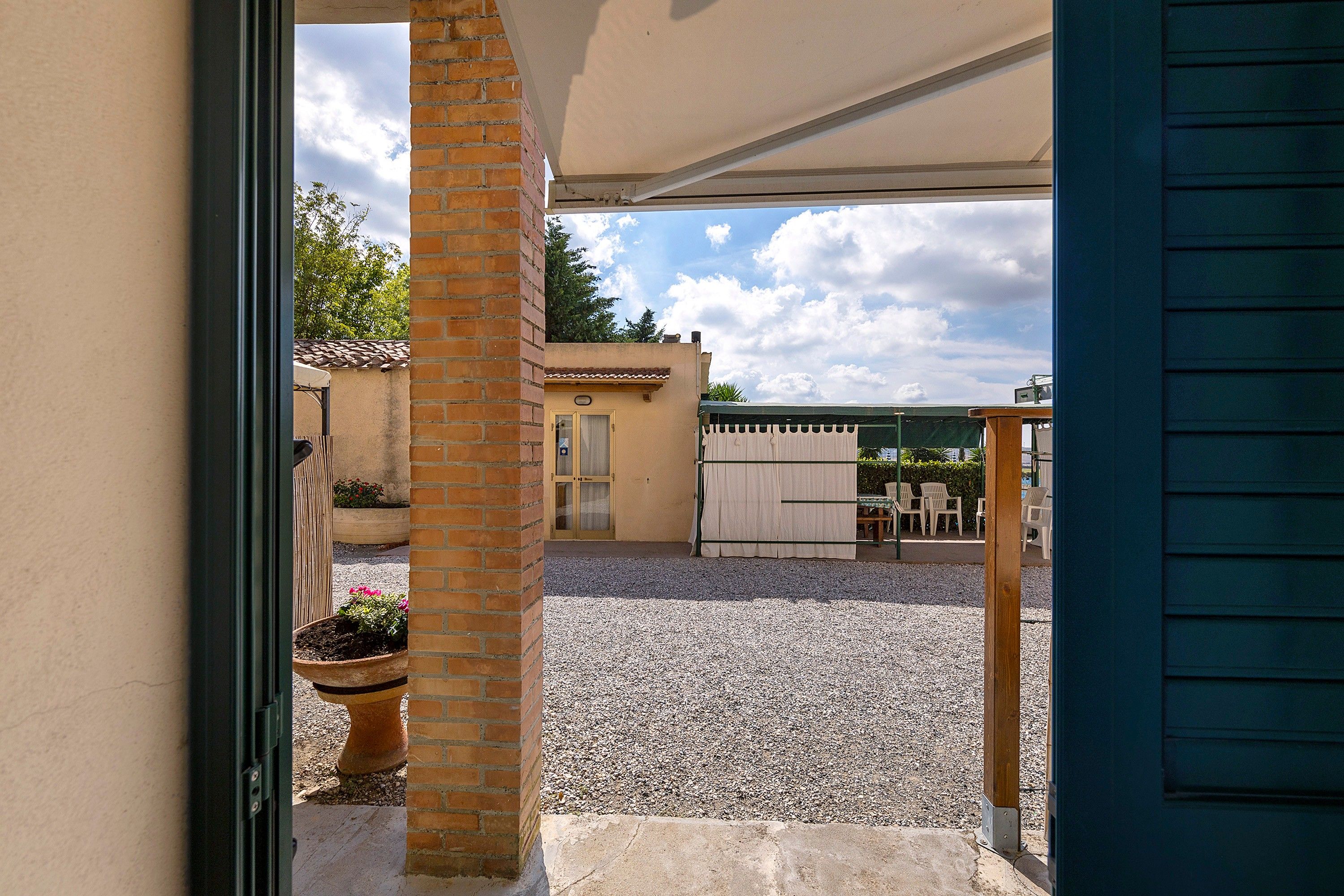 Eine überdachte Terrasse im Freien mit Kiesboden, einer Ziegelstütze und Blick auf ein Gebäude mit einem Ziegeldach.