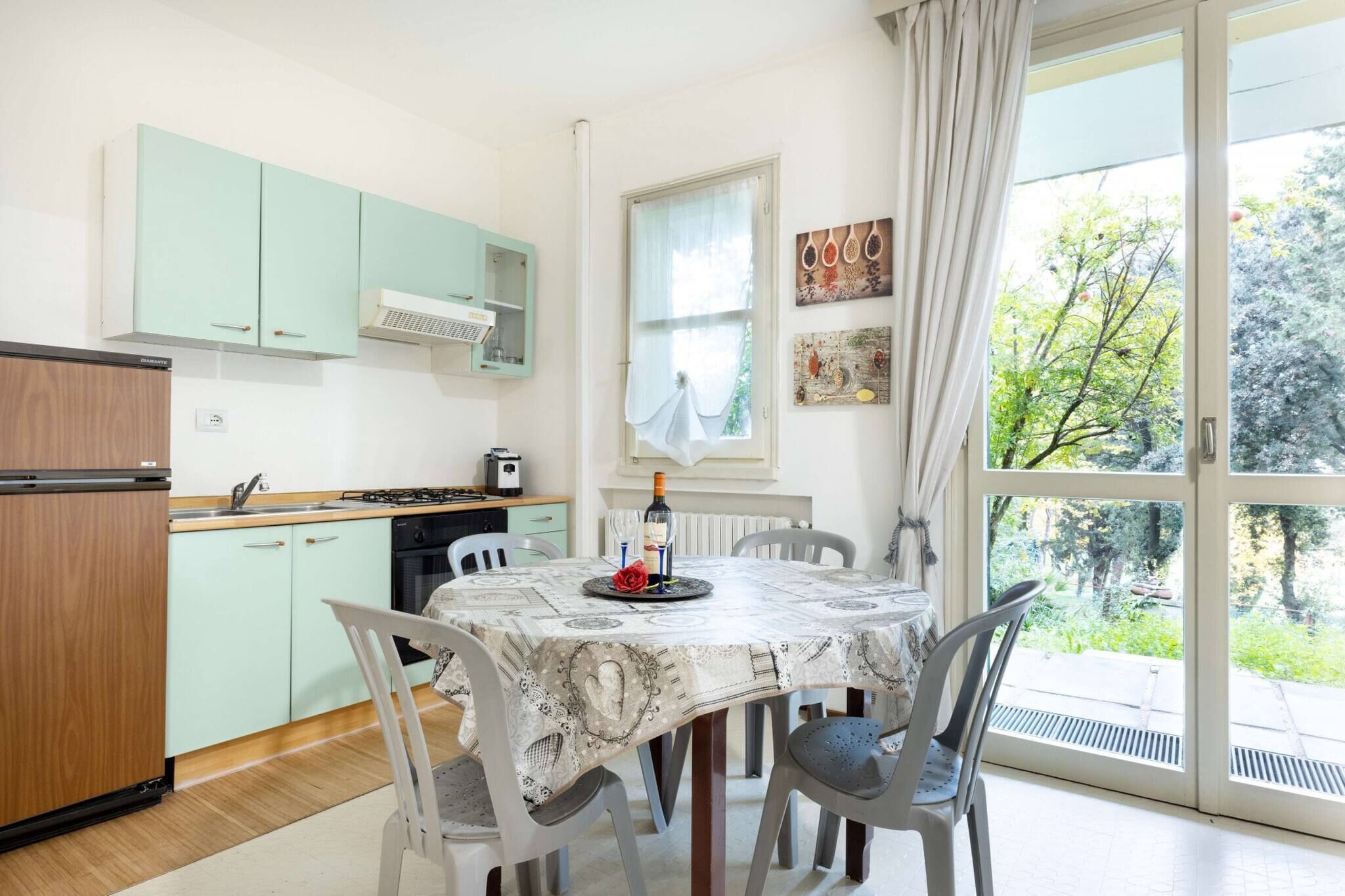 A kitchen with light green cabinets, a dining table with a patterned tablecloth, and a view of greenery through the window.