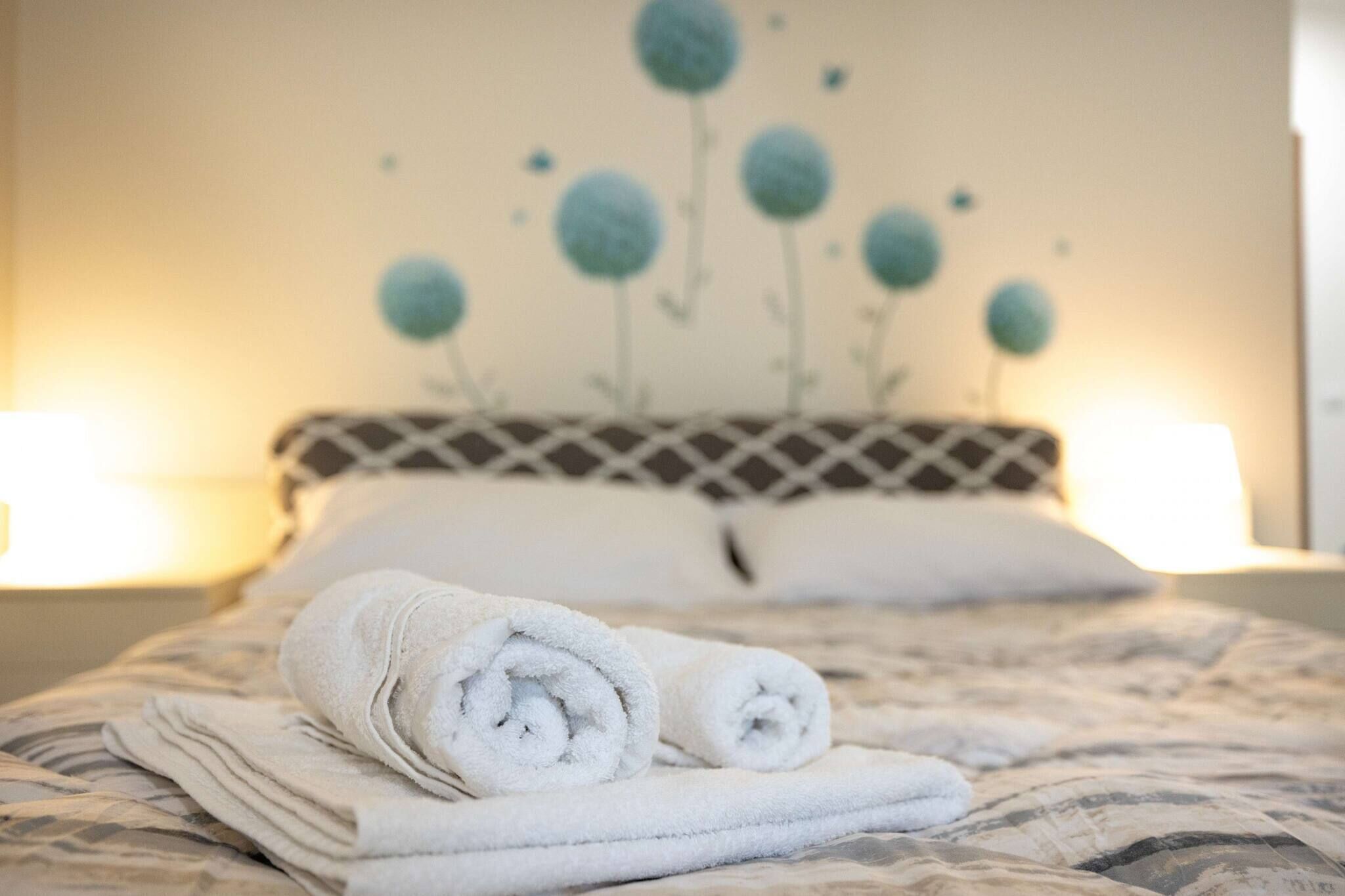 A neatly made bed with rolled white towels, a patterned bedspread, and a wall with a decorative floral design.