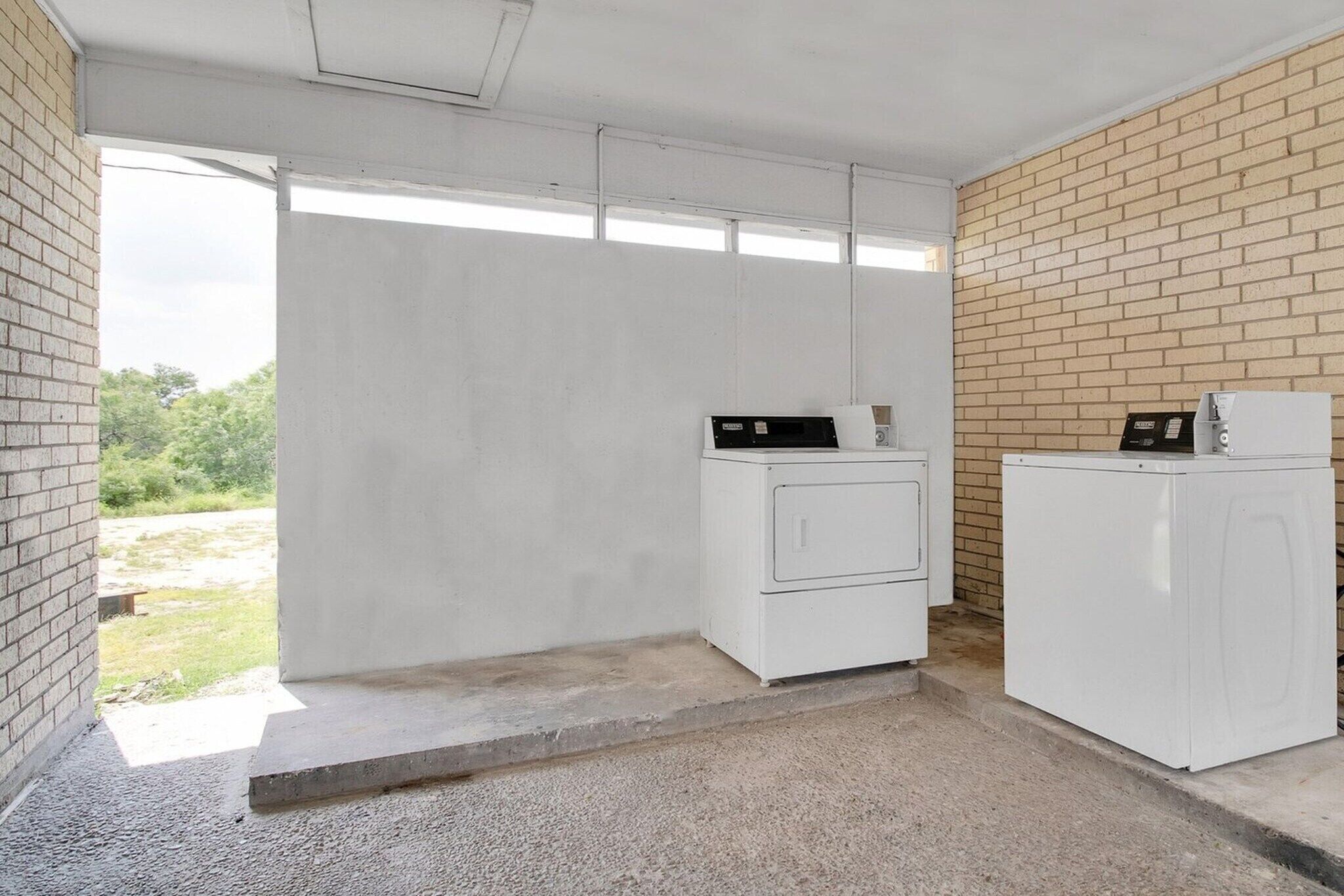 Una lavandería con dos lavadoras blancas, una pared de ladrillo y piso de concreto.