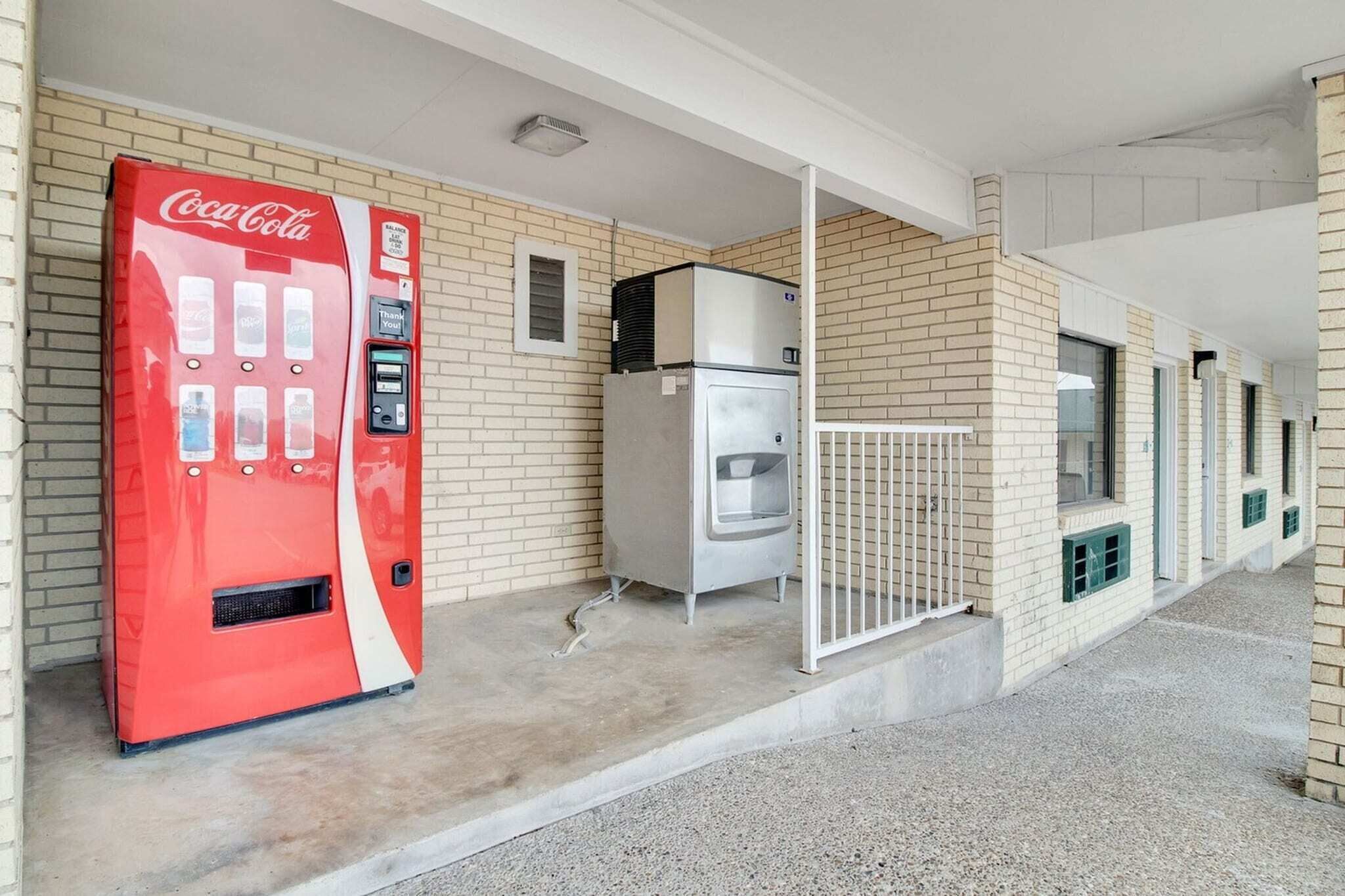 A Coca-Cola vending machine, a refrigerator, and a gated entrance to a building with a brick facade.